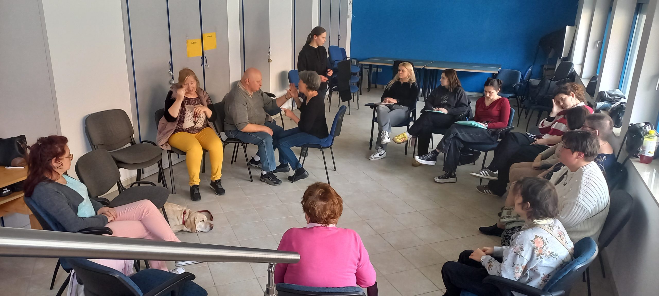 A közösségi nagy teremben körben ülnek a résztvevők, jobb oldalon két jeltolmács jelel, bal oldalon a tagok látszanak, majd mellettük gyógypedagógus hallgatók