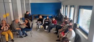 Szintén a félkörben ülő résztvevők látszanak és hallgatják a klubvezetőt aki a jeltolmácsok mellett áll