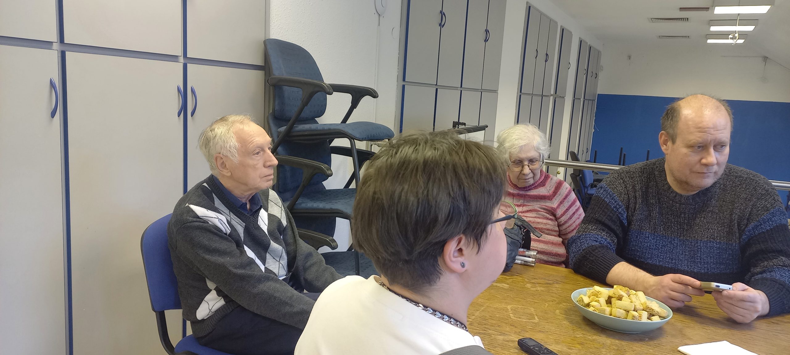 Csák Attila klubvezető és pár résztvevő egy asztal körül, a témaszemléltetéshez egy okostelefon, plusz sütemény az asztalon